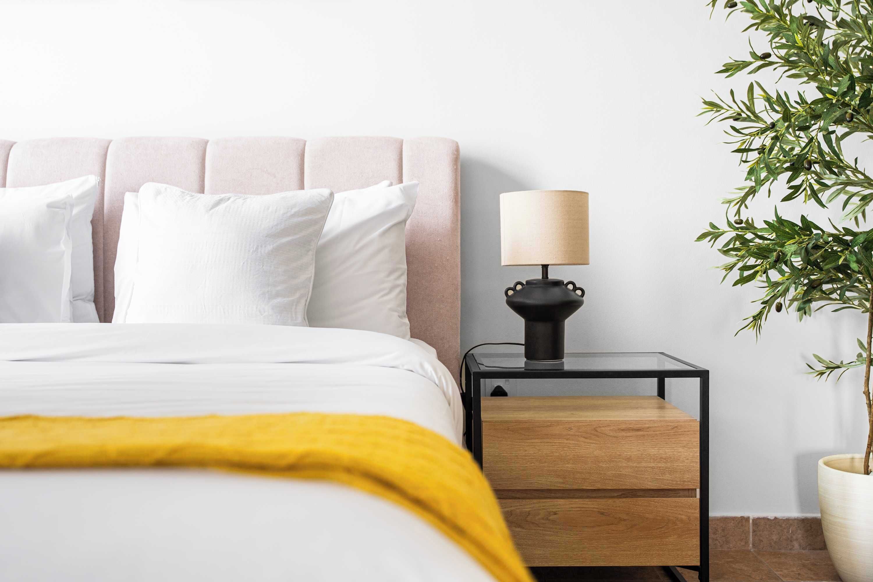 A bed with white bedding, a yellow throw, a bedside table with a lamp, and a potted plant.