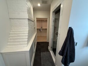 Een smalle gang met een witte, geribbelde wand, een glazen deur naar een badkamer, en een donker vloerkleed.
