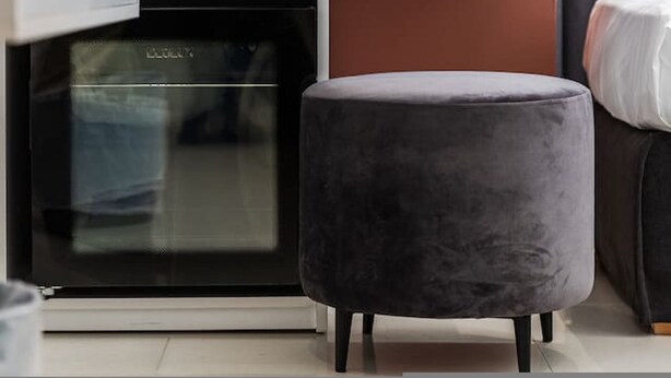 A compact kitchenette with a dishwasher, a small countertop, and a grey ottoman. - Brown Studio 116 Love in Colours Collection (Durrës)