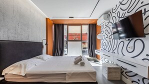 Quarto de hotel moderno com cama, televisão, secretária e uma janela grande com cortinas.