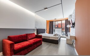 Een moderne hotelkamer met een rode bank, een bed, een bureau en een groot raam.