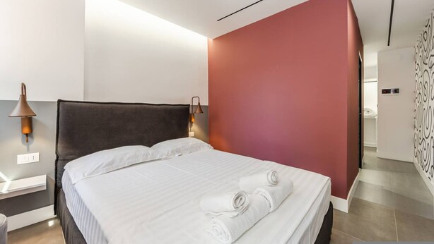 A modern hotel room with a bed, bedside lamps, a nightstand, and a wall with a geometric pattern. - Rose Studio 112 Love in Colours Collection (Durrës)