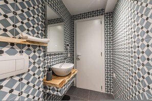 Ein Badezimmer mit einem Badezimmerboden in geometrischem Muster, einem weißen Waschbecken auf einem Holzboden, einem Spiegel über dem Waschbecken und einer weißen Tür.