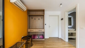 A room with a wooden bench, a small table, a wardrobe with hanging space, and a door leading to a bathroom. - Orange Studio 104 Love in Colours Collection (Durrës)