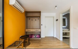 A room with a wooden bench, a small table, a wardrobe with hanging space, and a door leading to a bathroom. - Orange Studio 104 Love in Colours Collection (Durrës)