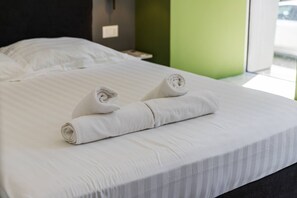 Cama bem arrumada com duas toalhas enroladas, uma mesinha de cabeceira e uma parede verde.