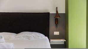 Ein ordentlich bezogenes Bett mit weißen Leinen, ein dunkles Kopfstück, eine Wandlampe, ein Nachttisch mit einem Buch und eine Steckdose.