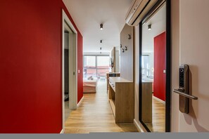 Un passadís amb una paret vermella, sòls de fusta i una porta corredissa de vidre que dona a una habitació amb un llit i un escriptori.