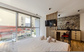 Habitació d'hotel moderna amb un llit gran, una televisió penjada en un panell de fusta, una cuineta petita i vistes a la ciutat a través de grans finestres.