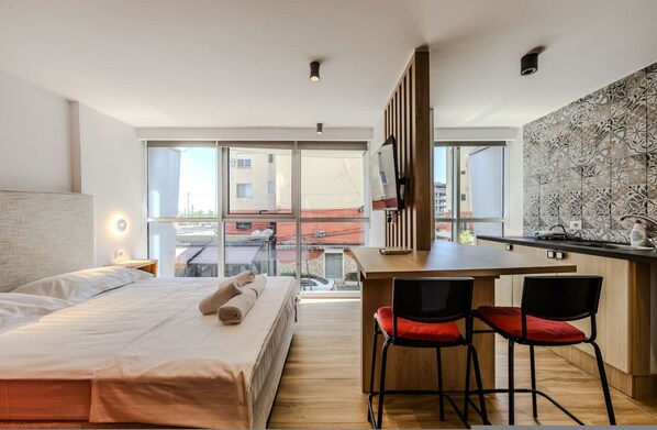 Habitació d'hotel moderna amb llit, escriptori amb dues cadires, zona de cuina i grans finestres que donen a una vista de la ciutat.