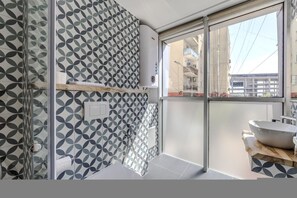 A modern bathroom with a geometric tile pattern, a glass shower enclosure, and a large window offering a view of the city. - Yellow Studio 106 Love in Colours Collection (Durrës)