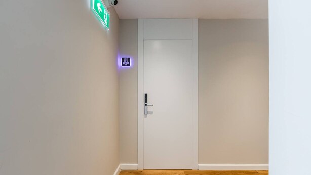 Un corridoio con una porta bianca, una segnaletica di emergenza verde e un piccolo pannello illuminato sopra la porta.