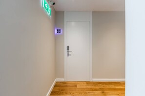 Um corredor com uma porta branca, um sinal de saída de emergência verde e um pequeno painel iluminado acima da porta.