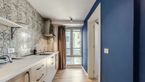 A narrow kitchen with patterned tiles, wooden cabinets, a window with curtains, and a blue wall. - Blue Studio 101 Love in Colours Collection (Durrës)