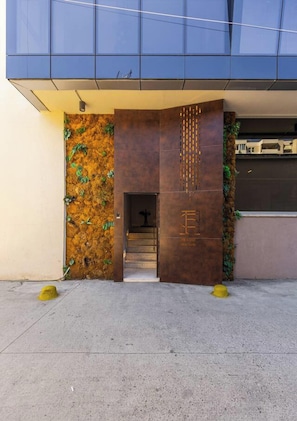 A modern building entrance with a glass facade, a textured wall with greenery, and a metal structure with a logo. - Olive Studio 105 Love in Colours Collection (Durrës)