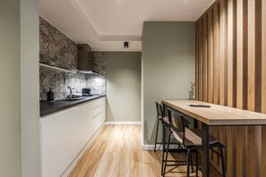 A compact kitchen with a wooden table and bar stools, a built-in stove, and patterned backsplash. - Olive Studio 105 Love in Colours Collection (Durrës)