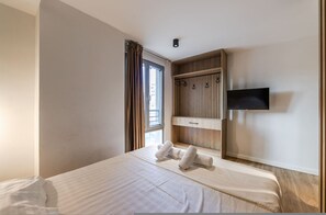A hotel room with a bed, a TV mounted on the wall, a wooden panel design, and a window with curtains. - Olive Studio 105 Love in Colours Collection (Durrës)