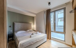 A modern hotel room with a large bed, a nightstand, a window with curtains, and a view of the outside. - Olive Studio 105 Love in Colours Collection (Durrës)