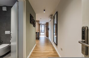 A modern hallway with a wooden floor, a bathroom on the left, and a door with a keypad lock on the right. - Olive Studio 105 Love in Colours Collection (Durrës)