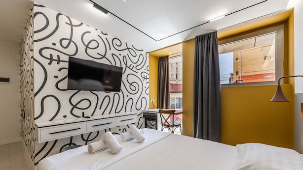 A modern hotel room with a large bed, a flat-screen TV mounted on a wall with a decorative pattern, a small seating area with a chair and table, and a window with curtains. - Golden Studio 117 Love in Colours Collection (Durrës)