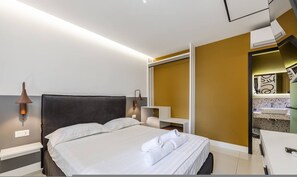 Quarto de hotel moderno com cama, abajures, escrivaninha embutida e banheiro com pia e espelho.