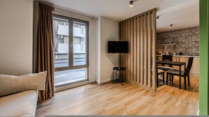A modern living room with a sofa, a television, a dining area with chairs, and a kitchen area with a countertop and cabinets. - Green Apartment 103 Love in Colours Collection (Durrës)