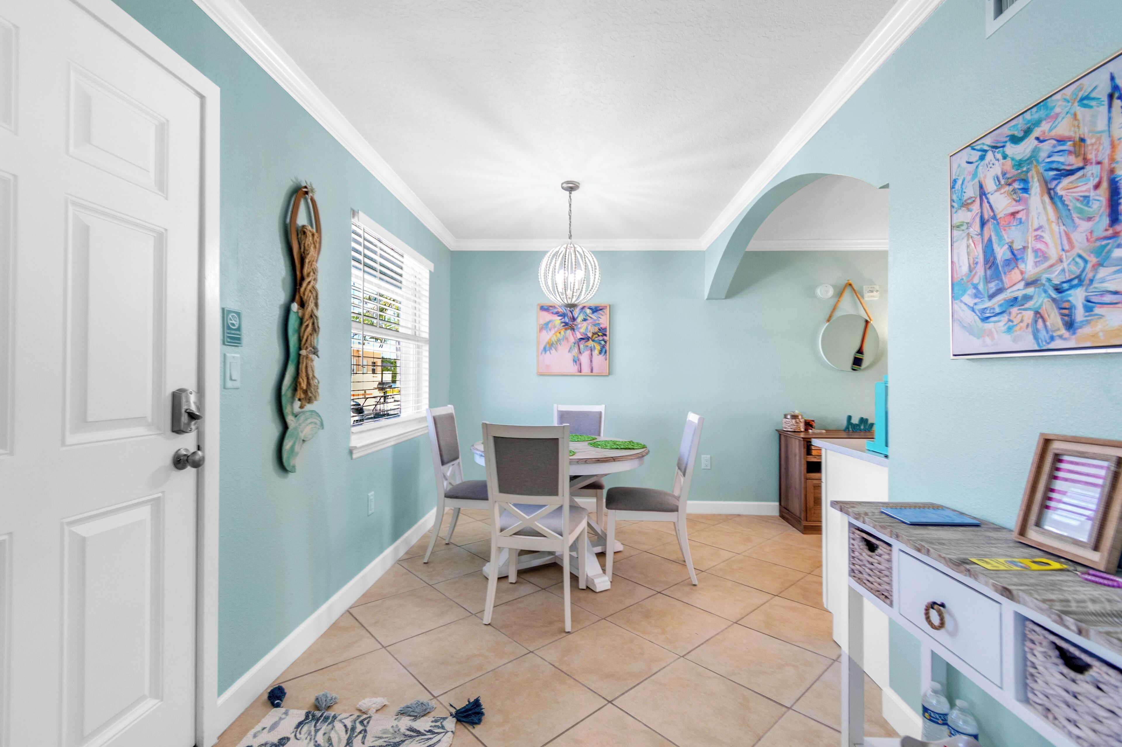 A dining area with a white table, chairs, and a chandelier. There is a painting on the wall and a small cabinet with items on top.