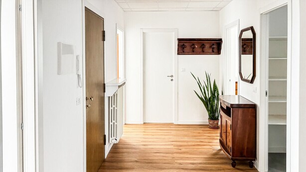Un couloir avec un plancher en bois, des murs blancs, une armoire en bois, une plante en pot et un miroir.