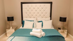 Una habitación de hotel con un cabecero capitonado, colcha color turquesa, almohadas blancas y dos lámparas de noche.