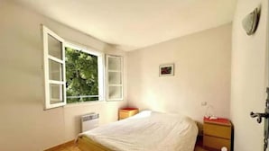 Un dormitorio con cama, ventana, radiador, mesita de noche y lámpara fijada a la pared.