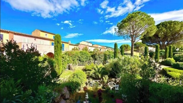 Un jardín con diversas plantas y árboles, una silla blanca y un edificio con fachada amarilla.