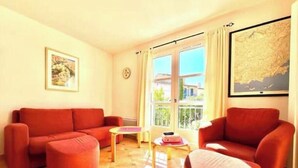 Una sala de estar con un sofá rojo, un reposapiés y un sillón, una mesita con libros y un ventanal con cortinas.