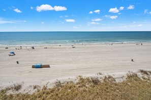 Ein weiter Sandstrand mit einigen Sonnenschirmen und Liegestühlen, einer Mülltonne und einem Rettungsschwimmerstand.