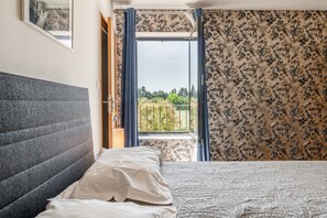 A bedroom with a grey headboard, a white bedspread, a window with blue curtains, and a balcony with a view of greenery.