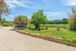 Une allée de gravier menant à une voiture garée, un mur de pierres et une pelouse bien entretenue agrémentée d’arbres et d’arbustes.