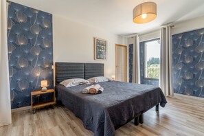 Quarto com cama, mesa de cabeceira, luminária e vista pela janela.