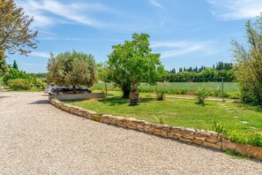 A gravel driveway leading to a parked car, a stone wall, and a well-maintained grassy area with trees and shrubs. - Bed and Breakfast 'Le Clapier' with Shared Pool, Wi-Fi and Air Conditioning (L'Isle-sur-la-Sorgue)