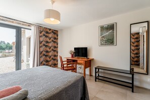 Una habitación de hotel con cama, escritorio con televisor, banco, ventana con cortinas e imagen enmarcada en la pared.