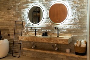 A bathroom with a stone wall, a white bathtub, a double sink with a mirror above, and a towel basket.