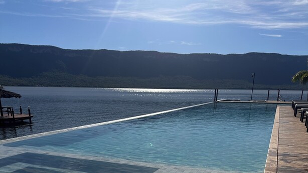 Una piscina infinita cristalina con una terraza de madera y vistas a una masa de agua y colinas lejanas.
