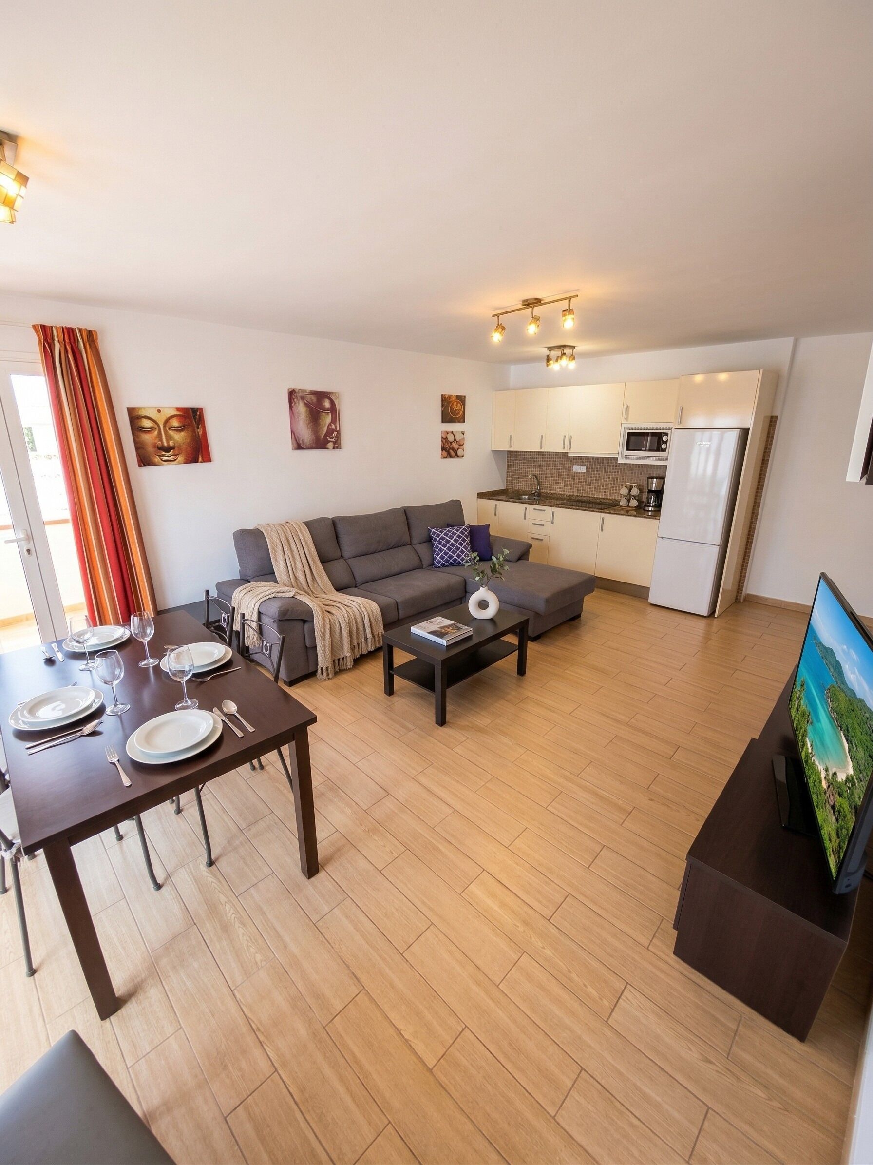 A modern apartment with a dining area, sofa, kitchen, and a TV displaying a scenic view.