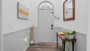 A hallway with a white door, a mirror, a painting, a vase, and a table with a plant. - Ideal Dream Getaway 4 Bedroom Home (Savannah)