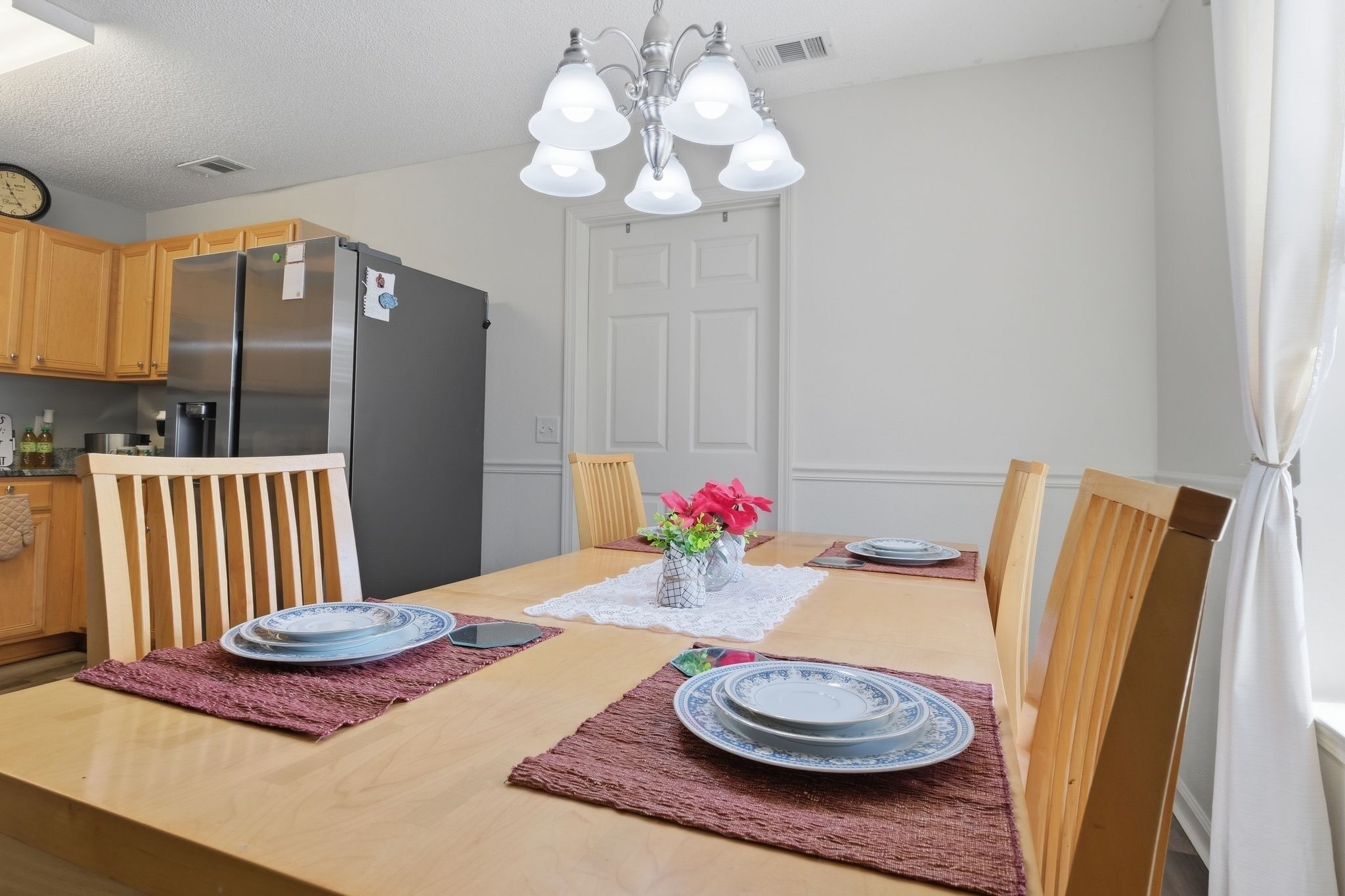 A dining area with a wooden table set for four, a refrigerator, wooden cabinets, and a chandelier.