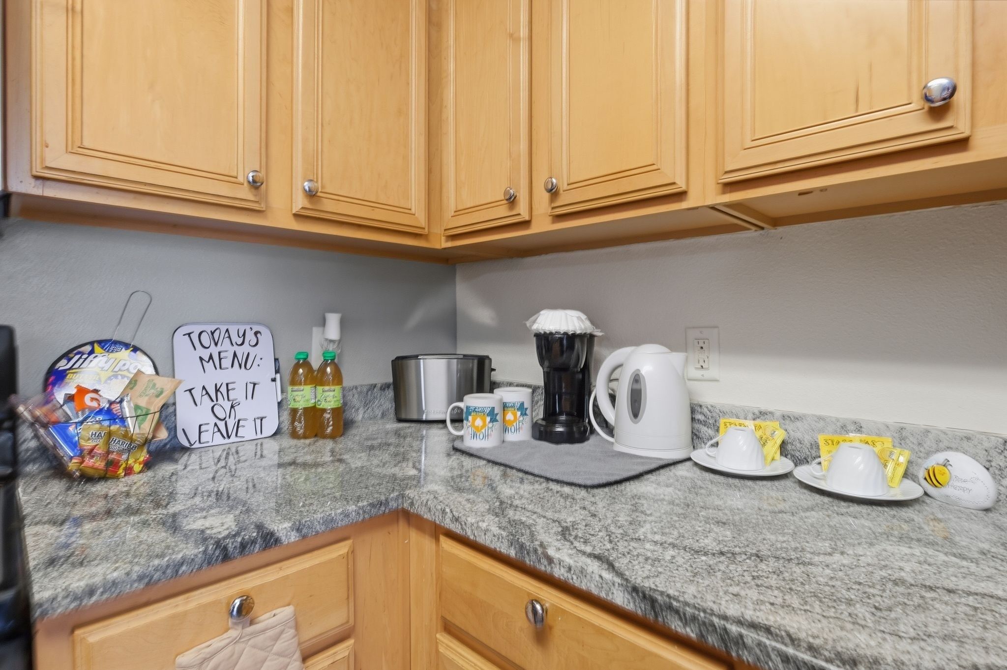 A kitchen with wooden cabinets, a granite countertop, a coffee maker, a kettle, and a sign with a menu.