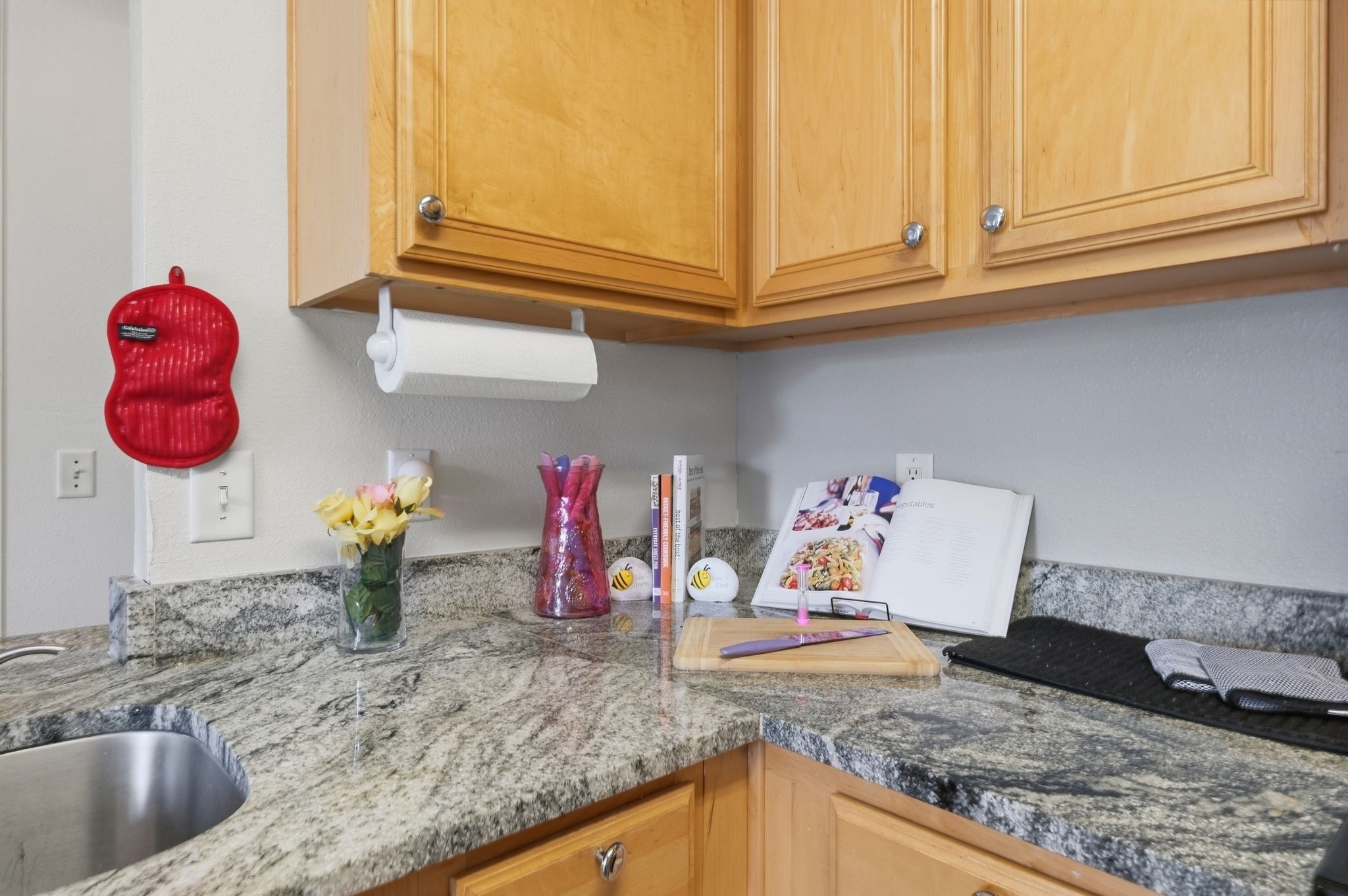 Une cuisine dotée de plans de travail en granit, de meubles en bois, d’un évier en acier inoxydable, d’un appareil électroménager rouge, d’un vase avec des fleurs et d’un livre de recettes.