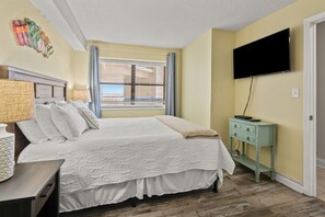 A bedroom with a bed, a bedside table with a lamp, a TV mounted on the wall, and a window with blinds. - Oceans 607 2 Bedroom Condo (North Myrtle Beach)