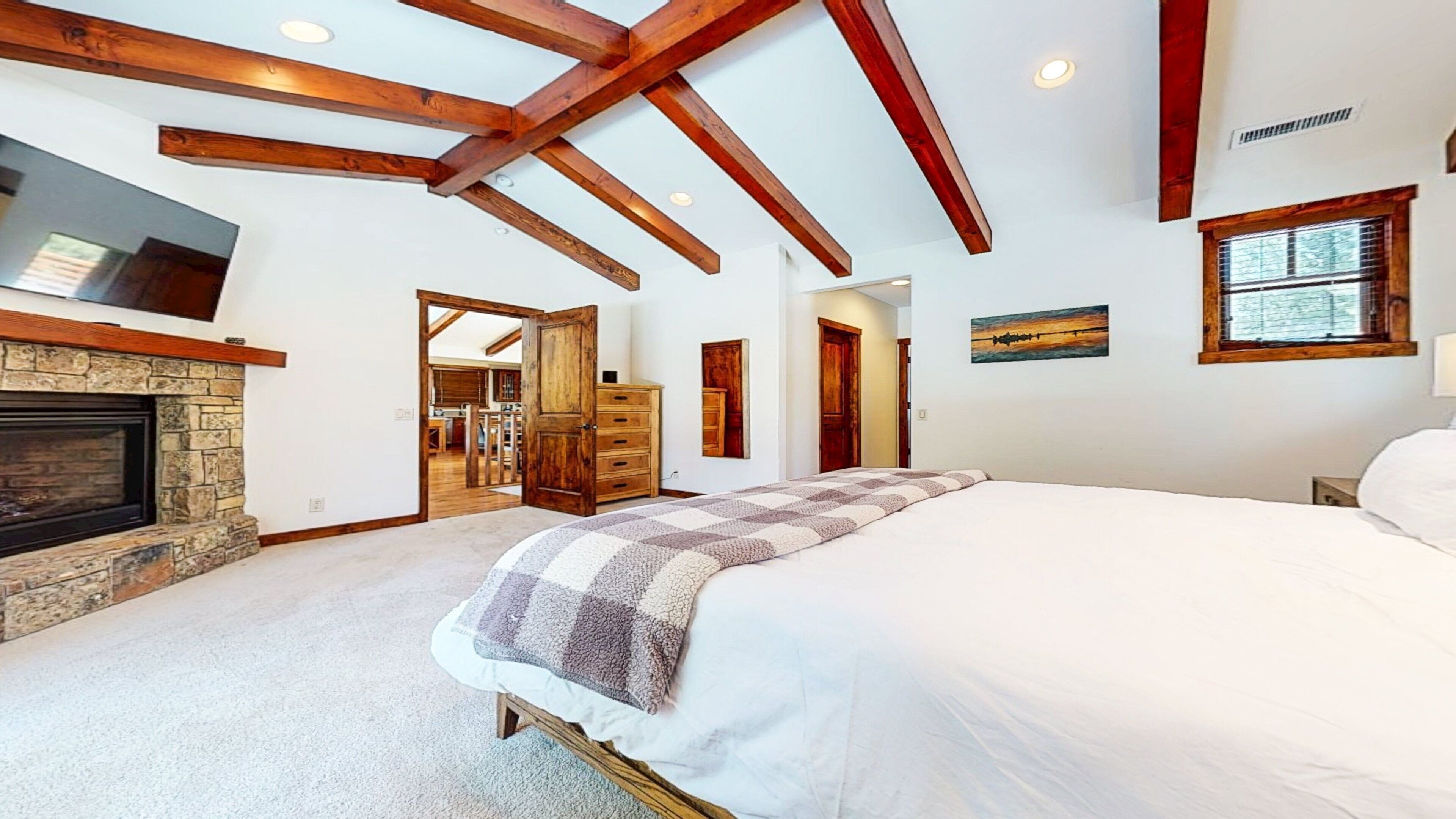 A bedroom with a large bed, a stone fireplace, wooden beams, a television, and a window.