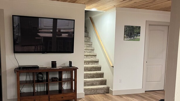 Een kamer met houten vloer, plafondventilator, televisie aan de muur, houten tv-meubel, trap en een deur.