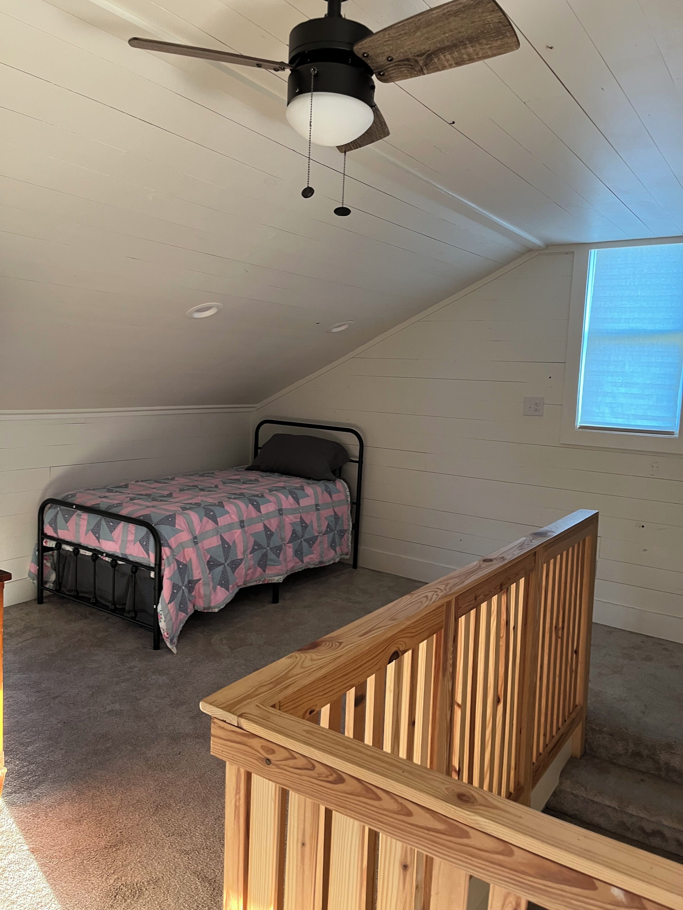 Ein Schlafzimmer mit schrägen Wänden, einem Bett mit gemustertem Überwurf, einem Hollaufstange und einem Deckenventilator.