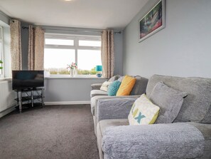 A living room with a grey sofa, a flat-screen TV on a stand, a window with curtains, and a painting on the wall. - Moorland View (St Austell)